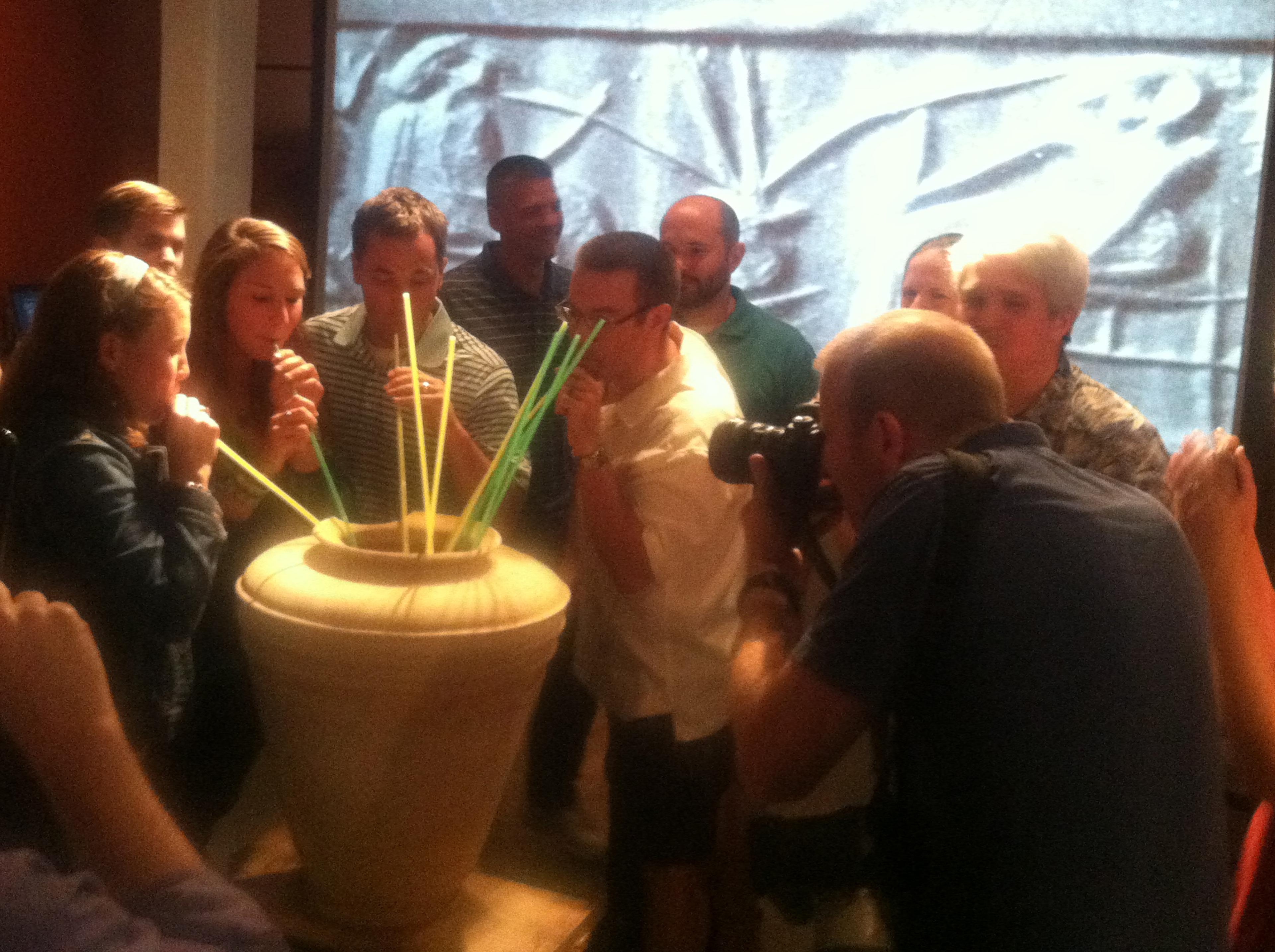 People drinking from a large pot using straws while a photographer captures the moment indoors.