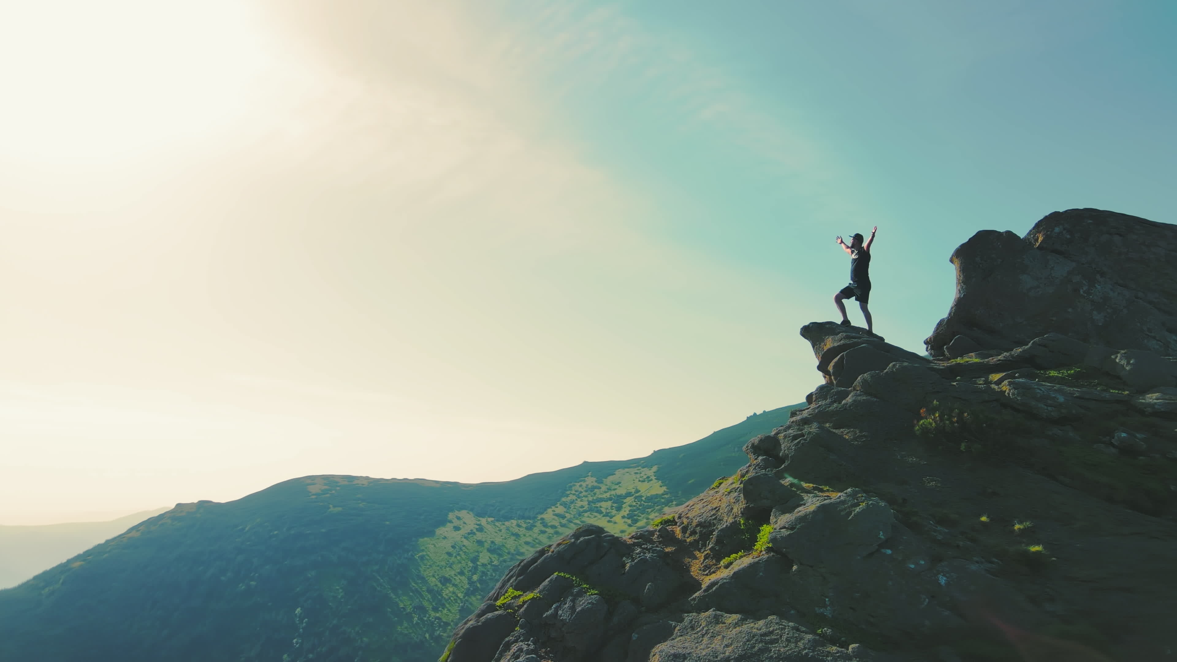 A man standing on top of a mountain with and raising his hands up the  mountain. Victory and overcoming the top. A tourist looking at the sunrise  on top of a rock.