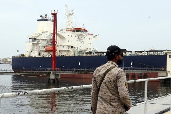 Чоловік у формі служби безпеки стоїть біля поруччя з видом на воду з великим синьо-біло-червоним нафтовим танкером позаду нього.