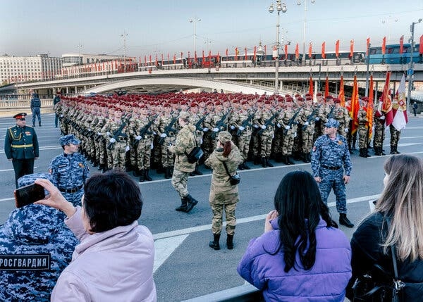 Російські солдати в формі стоять у строю на відкритому повітрі з глядачами на передньому плані та довгим мостом на задньому плані.