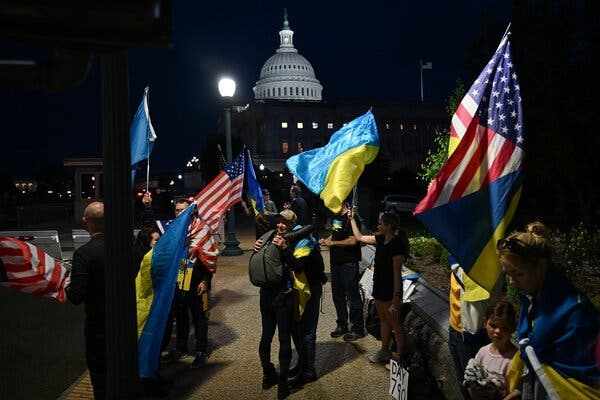 People waving Ukrainian and American flags at night, with the U.S. Capitol dome illuminated in the background.