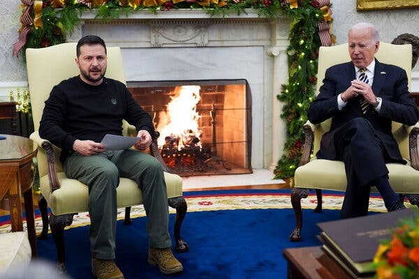 President Volodymyr Zelensky, in a dark sweater and green pants, sitting next to President Biden in front of a lit fireplace.