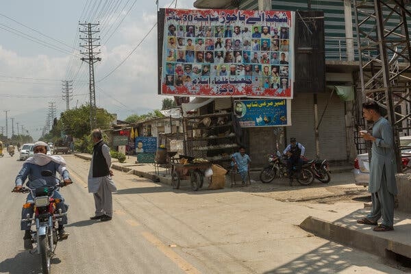 People standing at an intersection with a large billboard above it that shows many profile pictures.
