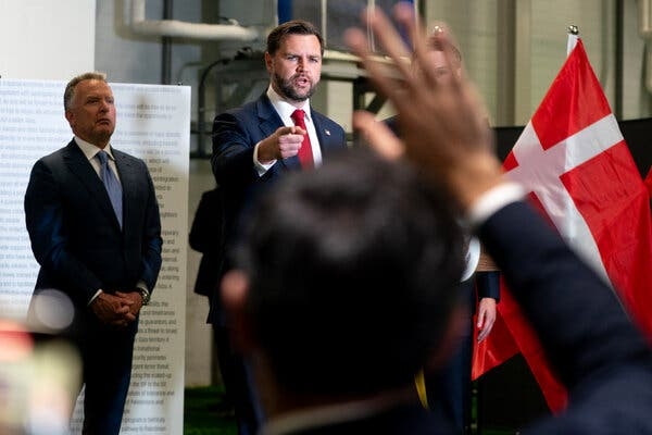 Steve Witkoff stands with his hands joined, while Vice President JD Vance points toward an audience out of frame. A member of the audience out of focus lifting his hand partly covers Jared Kushner, who stands by a red flag with a white cross.