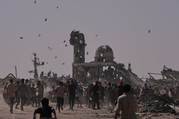 Leaflets float in the air as people are gathered under them. In the distance is a damaged building.