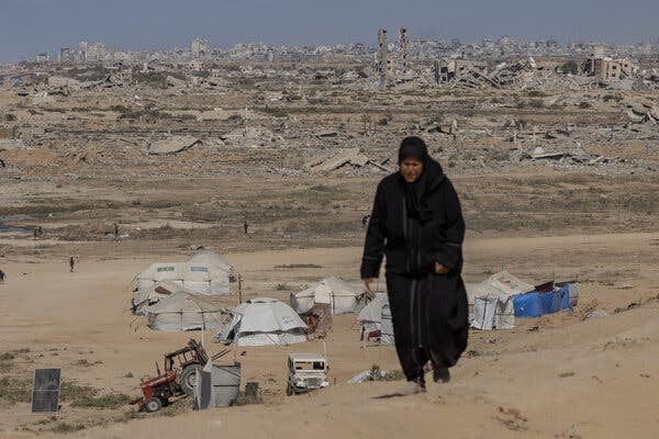 A woman walks in an arid landscape. Behind her are tents, parked vehicles and ruins.