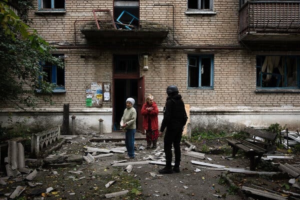 Three people, one wearing a black helmet and uniform, standing in front of a badly damaged apartment building.