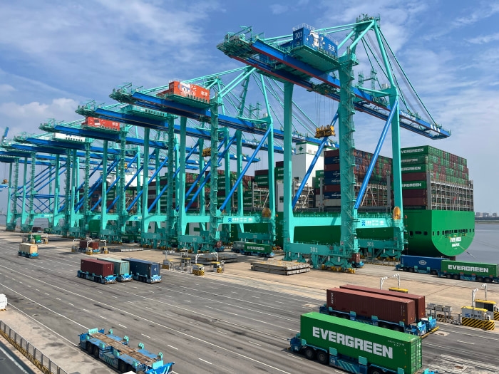 Container ship with Evergreen containers docked at Tianjin Port with multiple gantry cranes loading and unloading.