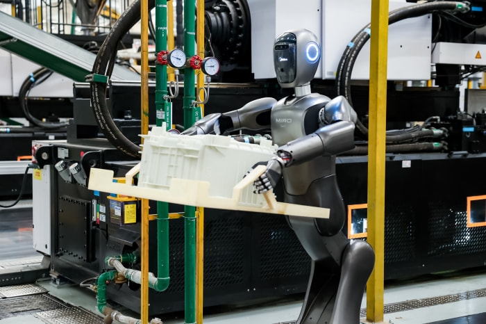 A gray Midea MIRO robot lifting a white crate on a wooden pallet in a factory.