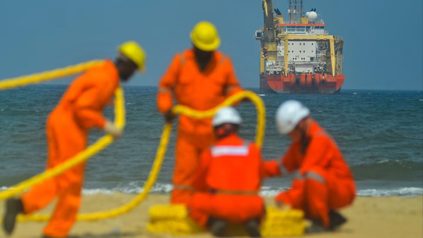 Workers in orange overalls and helmets handle yellow cable on a beach, with a large vessel offshore in the background