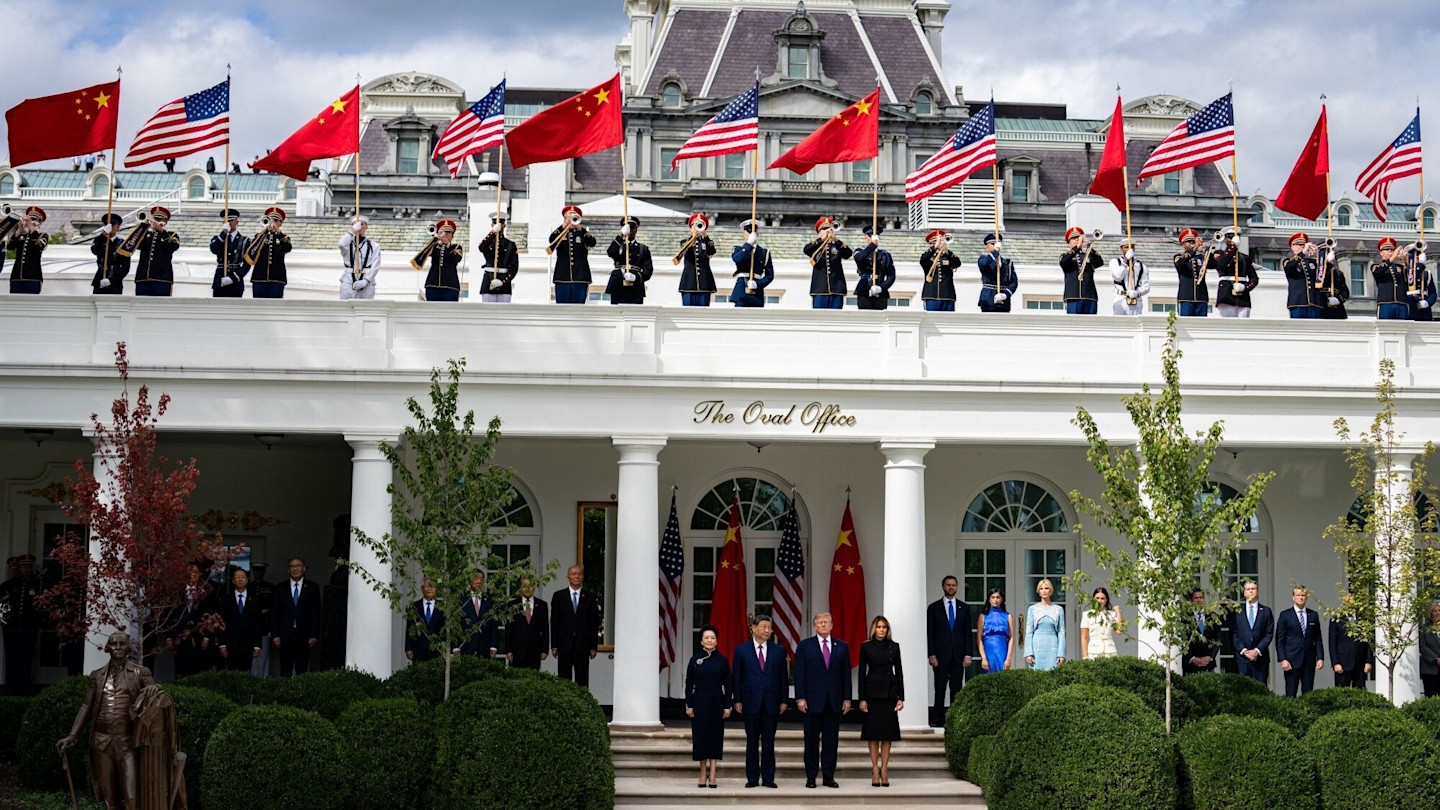 Peng Liyuan, Xi Jinping, Donald Trump, and Melania Trump stand together in front of the White House during a military review, with U.S. and Chinese flags displayed above.