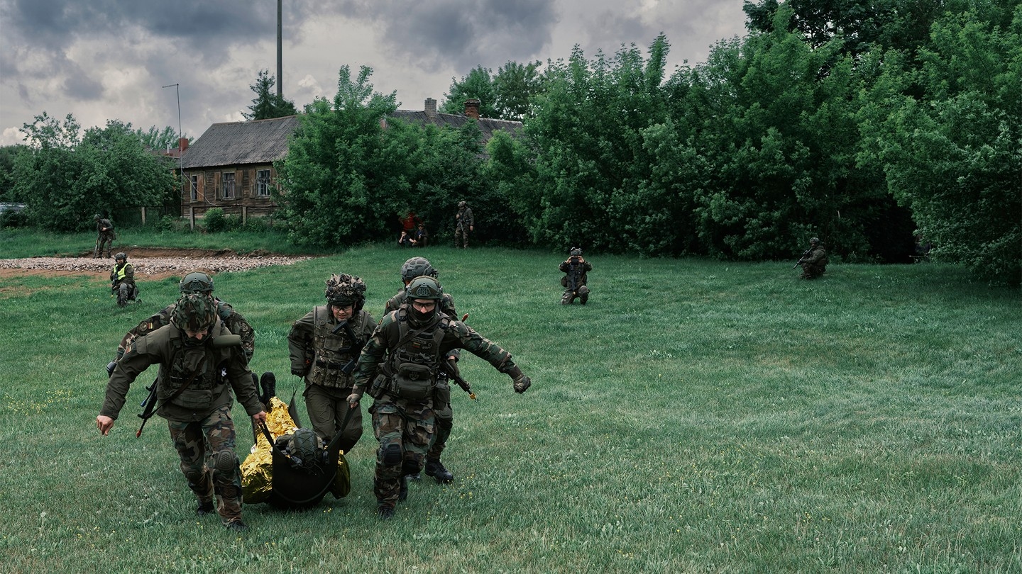Color photo of group of uniformed and heavily armed people running across green field carrying a person wrapped in gold thermal blanket with other militia members kneeling in background.