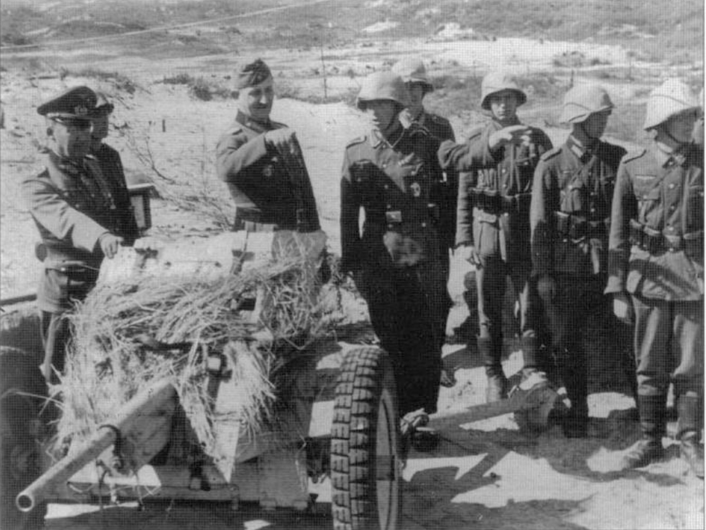 Два генерала оглядає позицію протитанкової гармати Pak 36 та її розрахунок в Тунісі. Вони всі споряджені шоломами з камуфляжними каверами з пішковини. 11 квітня 1943 року.