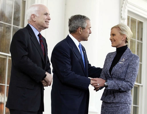 White-haired man in dark suit looks on as gray-haired man in dark suit holds hand and greets blonde-haired woman in medium-colored suit, all in front of a white building.
