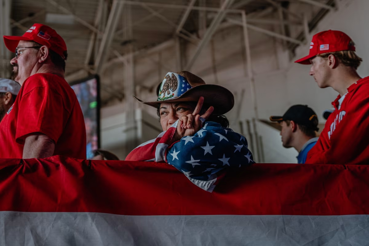 A woman sits wrapped in a Stars and Stripes flag. She is wearing a cowboy-style hat that shows part of the Mexican and US flags. Next to her stands a man in red shirt and red baseball cap