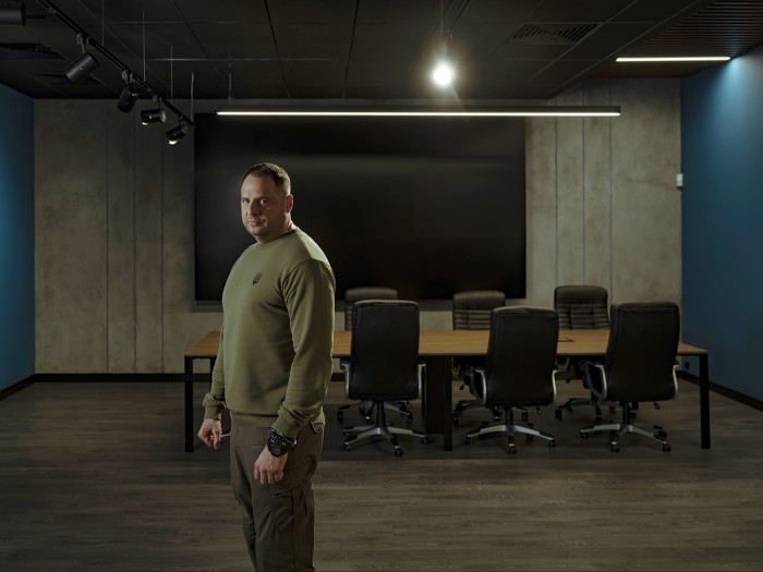 Man in a military-style sweatshirt stands in a dimly lit conference room