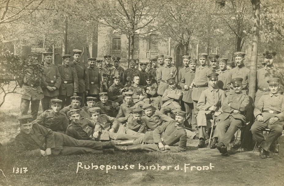sepia-toned archival photo of large group of uniformed WWI German soldiers, with a row standing, sitting, and lying down, and handwritten words 'Ruhepause hinter d. Front'
