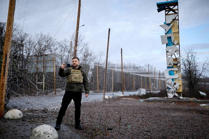 Volodymyr Zelenskiy records himself with a smartphone while standing near a damaged sign reading “Kupiansk.”