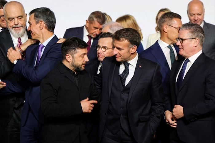 Volodymyr Zelenskiy gestures while standing next to Emmanuel Macron and Keir Starmer, who smile and look toward each other.