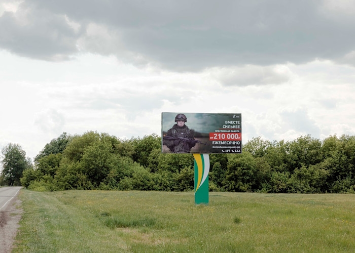 A Russian army recruitment billboard by a rural roadside shows a soldier in uniform with text advertising a monthly salary of 210,000 rubles.