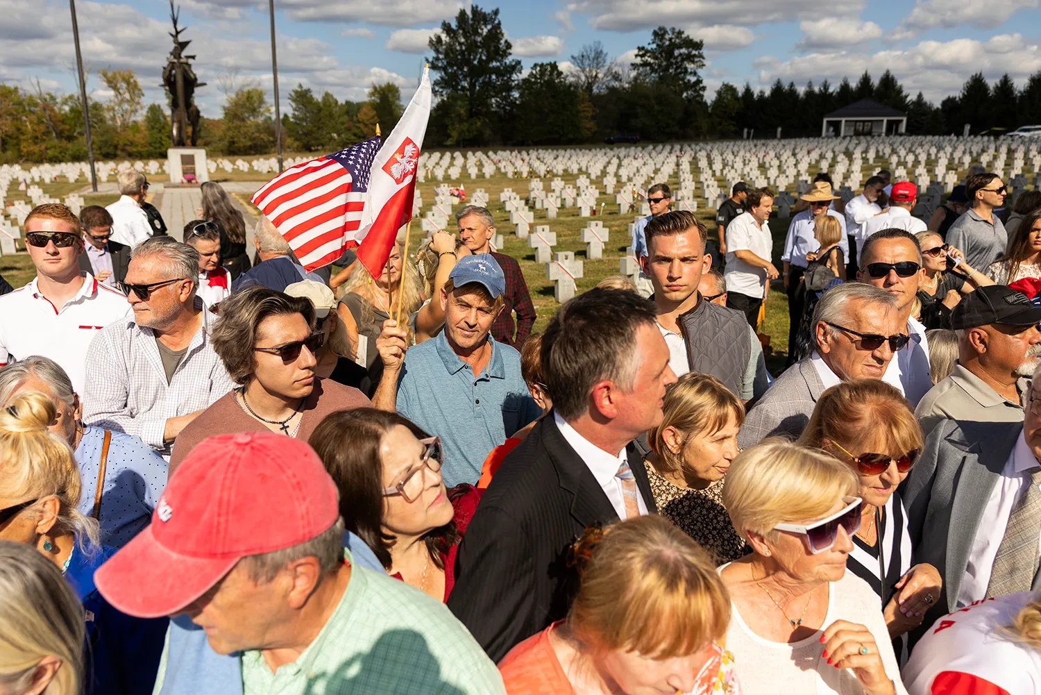 A crowd of people squint into the sun as they gather at a cemetery with rows of uniform-cross-shaped headstones visible in the distance. A U.S. and Polish flag are held by one attendee standing close the camera.