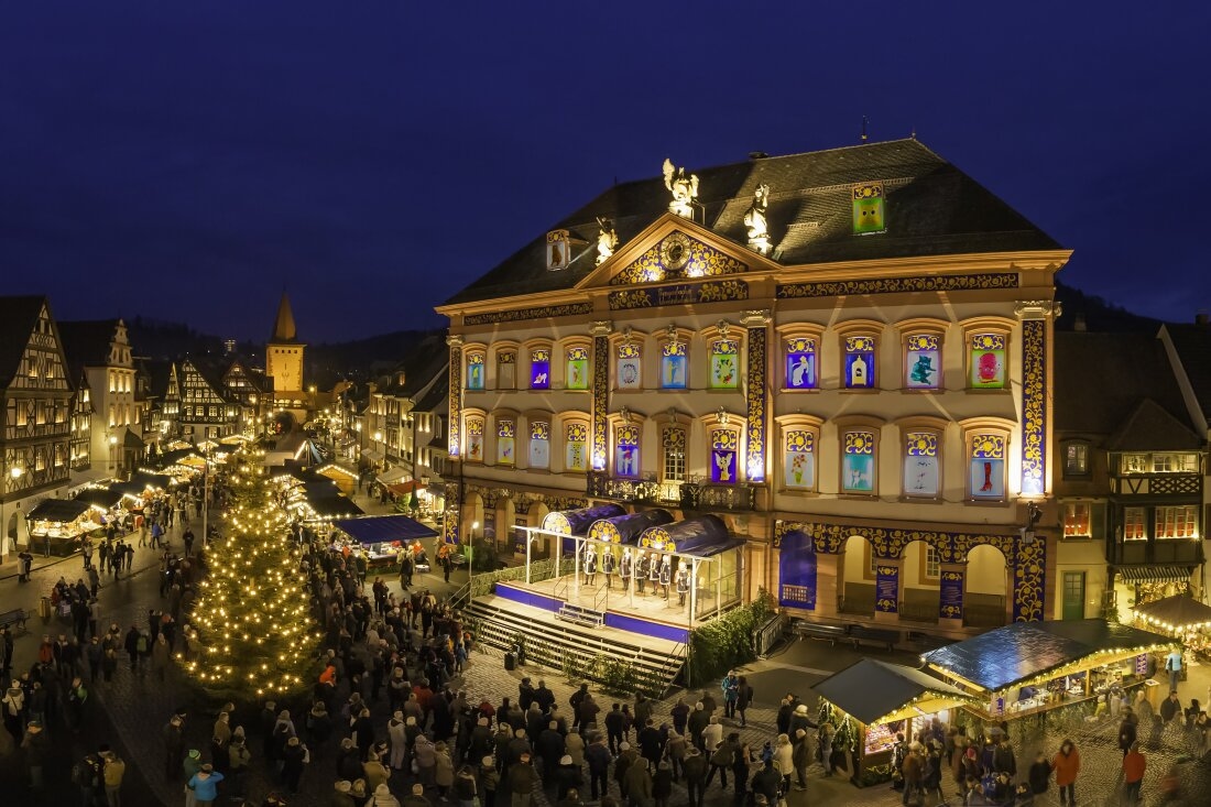 The town of Gengenbach, Germany, turns its town hall into a giant Advent calendar each December.  This photo shows the front of the neo-classical town hall at an angle, with each of the 24 window shades drawn up revealing a colorful image. It is evening. There is also a Christmas tree bedecked with lights and a Christmas market in the background.