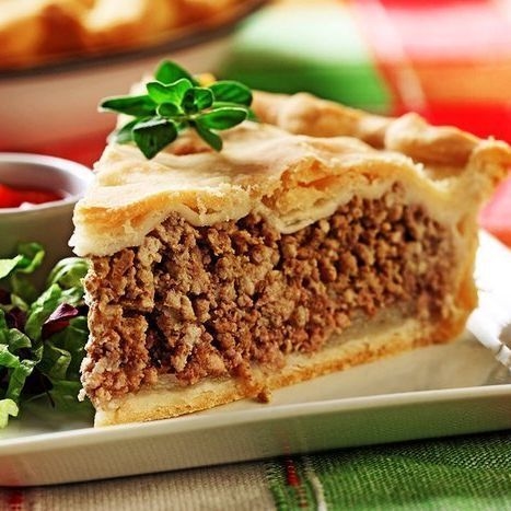 Close-up of a thick slice of tourtière (French-Canadian meat pie) on a white square plate. The golden flaky double crust is garnished with a sprig of fresh oregano. The filling is a savory mix of seasoned ground pork and beef. Beside the slice is a small garnish of green lettuce with purple tips and a ramekin of red ketchup. The background shows a blurred whole pie and a red-green plaid tablecloth. Warm, appetizing food styling.