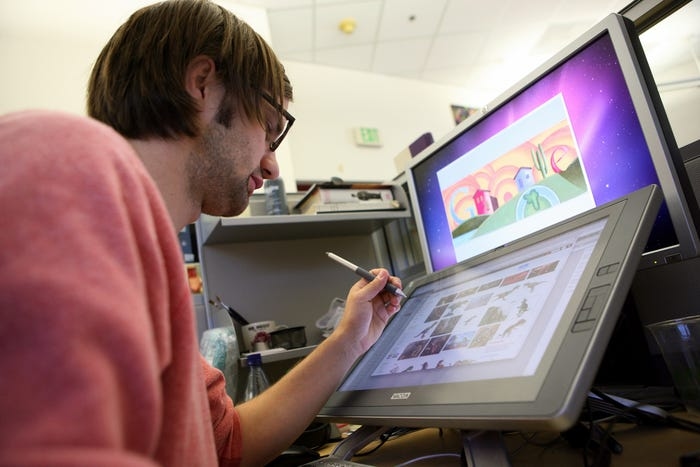 A Google employee sits in front of two screens, holding a stylus, while creating a Google Doodle illustration.